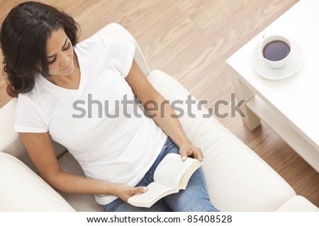 Beautiful happy young Latina Hispanic reading a paperback book and drinking tea or coffee at home on her sofa