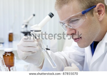 A male medical or scientific researcher or doctor looking at a test tube of clear liquid in a laboratory with microscopes.