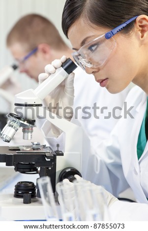 A Chinese Asian female medical or scientific researcher or doctor using a microscope in a laboratory with her colleague out of focus behind her.