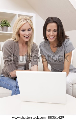 Two beautiful young women at home sitting on sofa or settee using a laptop computer and smiling