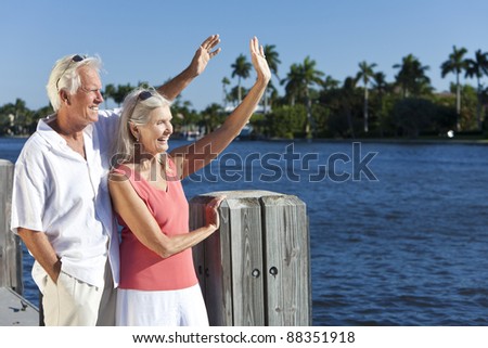 Happy senior man and woman couple together outside in sunshine waving by the sea on a jetty or pier