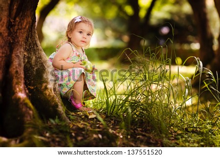 Cute baby girl eating apple under huge tree on green grass at sunny beautiful garden background