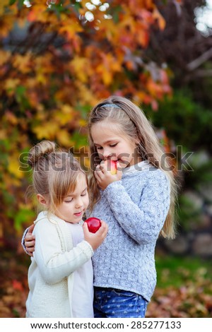Two cute little girls eating red apple outside at beautiful autumn background