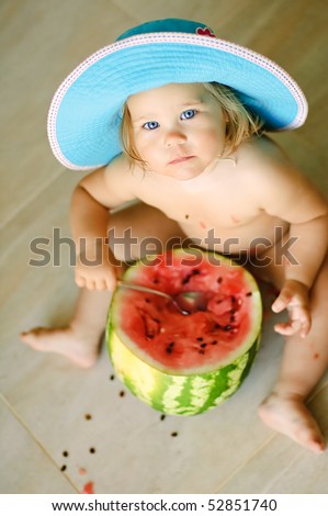 Cute baby-girl sits on a floor in a hat and eats a water-melon with a spoon