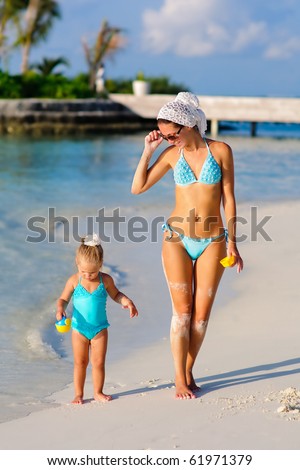 Young beautiful mother and her toddler pretty daughter in bikini walking on exotic beach