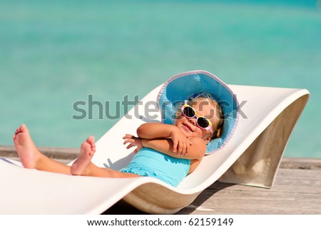 cute toddler girl in bikini, hat and sunglasses lyiing on a lounge and relaxing in tropic ocean background