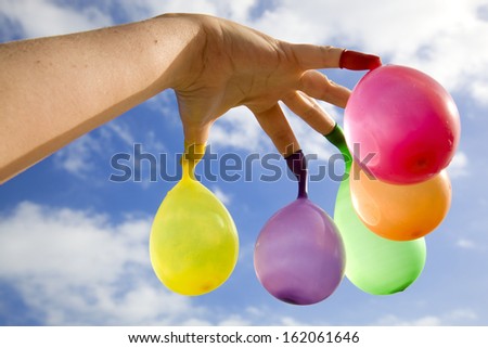 Open hand with multicolored water filled balloons hanging from the fingers, with a cloudy blue sky as background