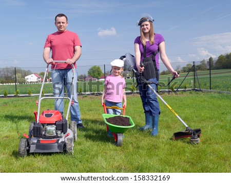Family gardening concept, dad standing with power mower, mom holding strimmer and their cute kid posing with green barrow loaded with soil