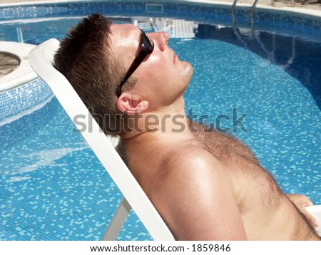 Young man in sunglasses laying on a deckchair sunbathing by the swimming pool