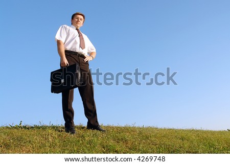 Confident businessman in white shirt and tie standing on grass with black briefcase against clear blue sky