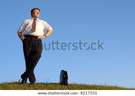 Businessman in white shirt and tie standing with crossed legs in grass field against clear blue sky looking ahead