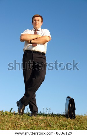 Businessman in white shirt and tie standing with crossed legs in grass field against clear blue sky with confident look