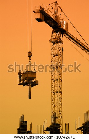 Silhouette of construction worker being transported by jib crane with concrete mix charging hopper against sunset sky