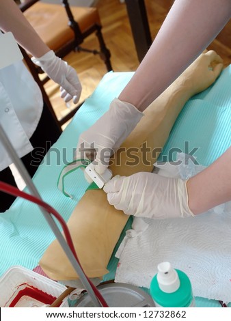 Nursing student practicing blood taking on mannequin hand under instructor supervision - putting compression band