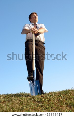 Businessman in white shirt standing in grass field over clear blue sky with one leg resting on shovel driven into grass looking ahead - new business vision concept