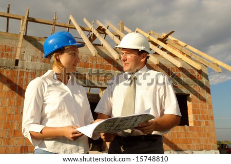 Female and male building designers discussing building plans against unfinished brick house with wooden roof structure