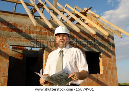 Building engineer wearing white helmet  holding building plans standing over unfinished brick house with wooden roof structure