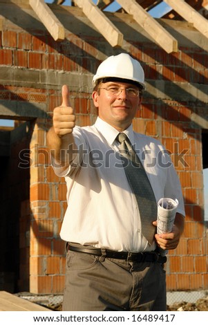 Civil engineer wearing white helmet holding roll of building plans showing thumb up sign over unfinished house