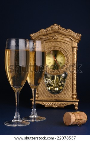 Two champagne glasses, champagne cork and table clock showing midnight over dark blue background