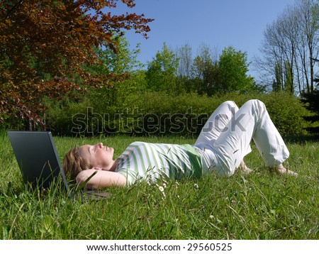 Young woman lying on back on grass with dreamy look with laptop placed by her head