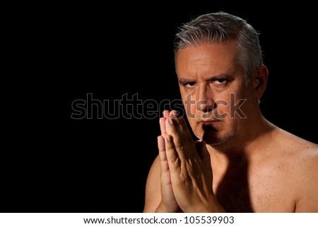 Handsome unshaven middle age man with salt and pepper hair and bare chested with hands together in a prayer pose on a black background.