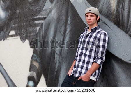 Handsome young man in an urban fashion lifestyle pose leaning on a graffiti wall wearing a baseball cap.