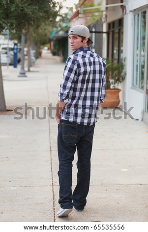 Handsome young man in an urban fashion lifestyle pose walking down a sidewalk along store fronts and looking back.