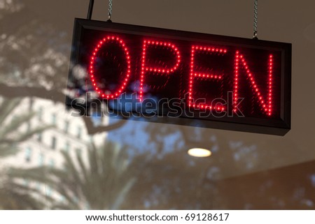 Store open sign with window reflections of a downtown scene with trees, building, and blue sky in the background.