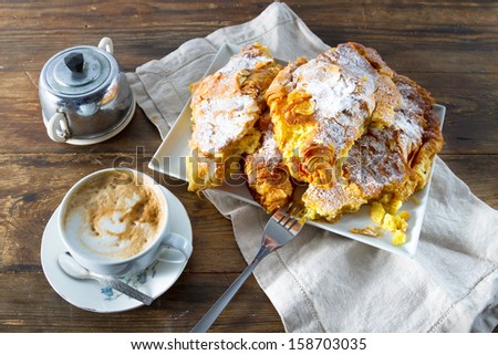 Continental breakfast: delicious almond croissants sprinkled with icing sugar and with a custard filling, accompanied by a cappuccino, on a rustic wooden background.