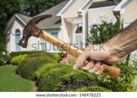 Man\'s hand holding hammer in front of a house indicating home improvement and maintenance.