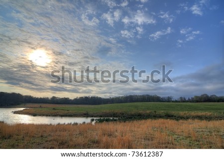 River winding through a field with the sun in the sky behind busy clouds.