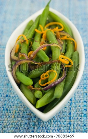 Green beans with caramelized red onion and orange zest in white boat shaped serving dish on blue textured background, vertical format