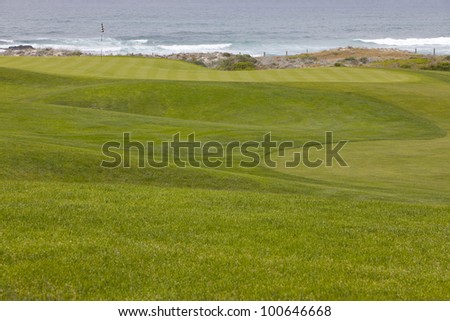 Beautiful, manicured golf course greens on a softly rolling hilly topography leading to the hole. The putting green is located right by the ocean, with small waves washing into shore.