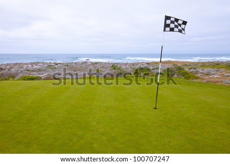 Beautiful putting green and pin flag fluttering in the ocean breeze on a scenic, golf course situated on the Pacific Ocean. Dramatic cloudy sky overhead.
