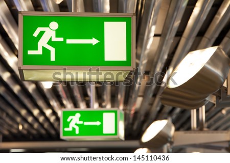 Illuminated Green Exit Signs Attached To The Ceiling In A Public Transportation Facility. Signage Of A Human Figure Running And Exiting Through A Door Opening. Lights Shine On Ceiling And The Signs.