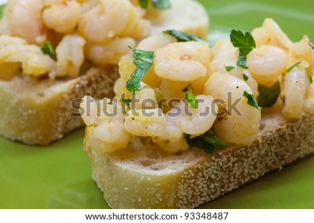Small shrimp dressed in a garlic, red pepper flake, ground pepper, and curry dressing presented on a slice of sourdough bread. Shrimp tapas presented on a green plate.