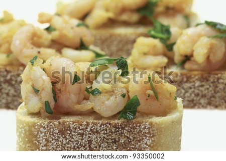 Small shrimp dressed in a garlic, red pepper flake, ground pepper, and curry dressing presented on a slice of sourdough bread. Shrimp tapas presented on a white background.
