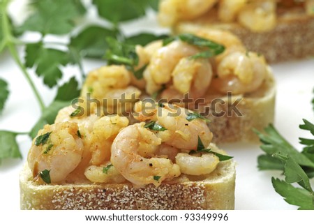 Small shrimp dressed in a garlic, red pepper flake, ground pepper, and curry dressing presented on a slice of sourdough bread. Shrimp tapas presented on a white background garnished with parsley.