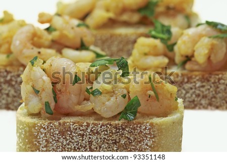 Small shrimp dressed in a garlic, red pepper flake, ground pepper, and curry dressing presented on a slice of sourdough bread. Shrimp tapas presented on a white background.
