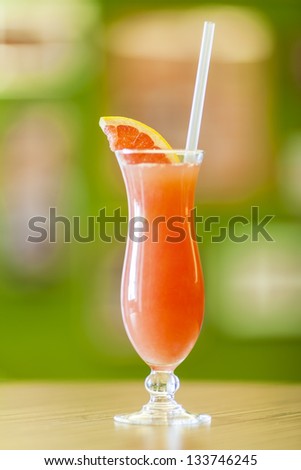 Freshly squeezed grapefruit juice served in a hurricane glass with straw and slice of grapefruit on blurred green background and brown desk