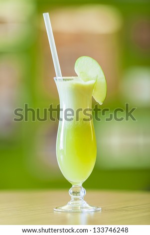 Freshly squeezed apple juice served in a hurricane glass with straw and slice of apple on blurred green background and brown desk