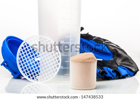 Scoop of whey protein in front of gym equipment: gloves and shaker parts