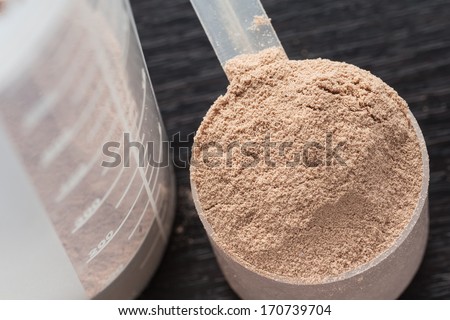 Scope of chocolate whey isolate protein next to the translucent protein shaker, with focus on the protein inside the scoop