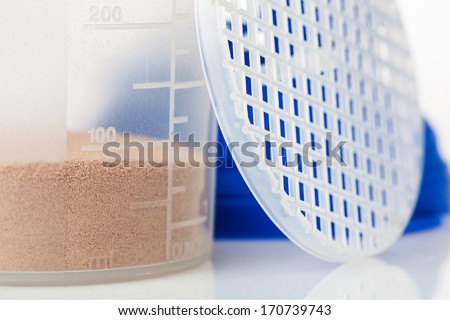 Closeup of chocolate whey isolate protein inside the shaker, with protein shaker parts leaning against the shaker, with focus on number 100 on the measure, on white background