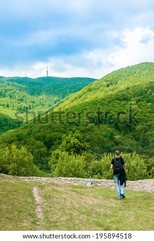 Unidentified photographer carrying a tripod and walking towards the rock wall preparing to take a photo of sunlit wood on the hill, with TV tower on the top of the hill