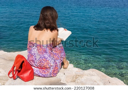 Young female reading a book on the rocky beach by the sea in the Adriatic