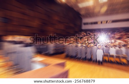 Audience in big concert hall during graduation ceremony. Camera flash in crowd goes off. Concept of graduation ceremony. Radial zoom effect defocusing filter applied, with vintage instagram look.