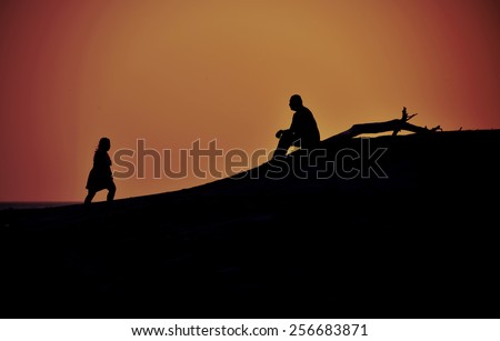 Silhouettes of a woman walking towards a man that is sitting at a beach during  sunset