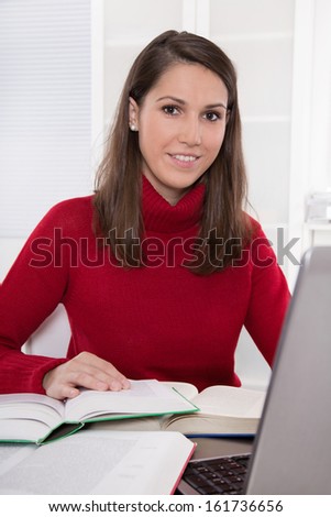 Reading and research: brunette woman sitting in red jumper at desk with books and laptop