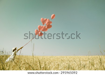 Young romantic girl with red heart balloons walking in a field of wheat.